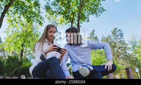 Two high school students are sitting on a bench in a park, looking at a smartphone and enjoying their school day Stock Photo