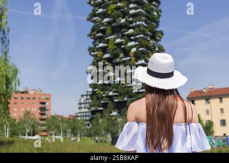 Girl tourist with a sun hat looking at a skyscraper with plant and tree-covered balconies and terraces. Architecture, travel, and lifestyle concepts. Stock Photo