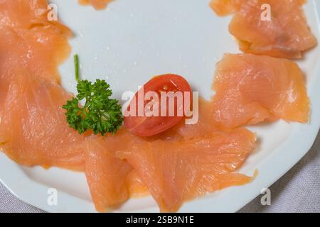 A close-up of delicately arranged smoked salmon slices on a white plate, complemented by a fresh sprig of parsley and a halved cherry tomato. Stock Photo