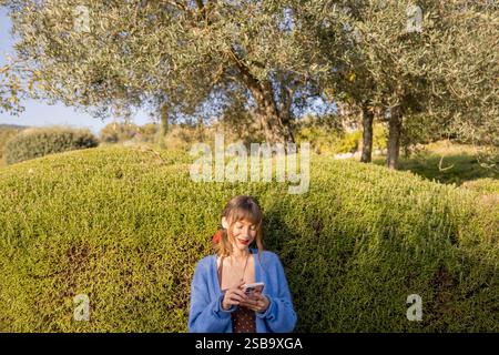 Smiling woman in a blue cardigan using a smartphone, standing next to lush green hedges with olive trees in the background under a bright sunny sky Stock Photo