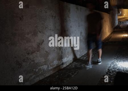 A blurred figure walks on a dimly lit path next to a textured concrete wall at night. Stock Photo