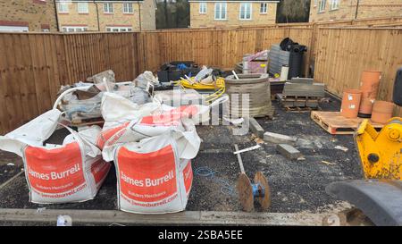 Construction site materials are scattered within a fenced area.  Two large builder's bags, various building blocks, pipes, and a small excavator are v Stock Photo