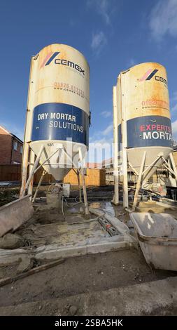 Two weathered Cemex Ready Mix dry mortar silos stand on a construction site.  They are supported by metal frames, and surrounded by earth, building ma Stock Photo