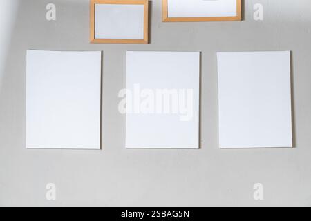 Three blank white sheets of paper arranged on a light gray wall, with two empty wooden frames above them. Stock Photo