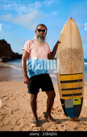 Senior man with grey beard stands on sandy beach holding surfboard. Elderly surfer enjoys sunny day by ocean. Retirement active lifestyle and wellness Stock Photo