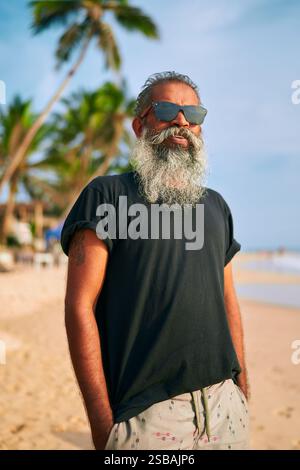 Senior bearded man in sunglasses relaxes on sunny tropical beach. Elegant older male enjoys warm weather, ocean breeze. Stylish traveler in casual Stock Photo
