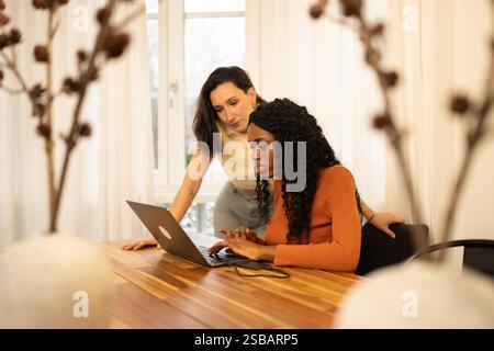 Two diverse businesswomen are working together on a laptop in a modern office, demonstrating teamwork and collaboration in a professional setting with Stock Photo