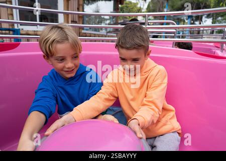 Two cheerful boys enjoying a ride on a spinning carousel at an amusement park, creating joyful childhood memories Stock Photo