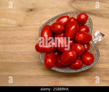 A clear glass bowl filled with vibrant red cherry tomatoes is placed on a rustic wooden cutting board. Perfect for salads or simple snacks, these toma Stock Photo