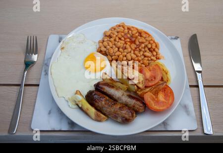 Traditional English breakfast featuring eggs, sausages, baked beans, tomatoes, and potatoes on a white plate. Stock Photo