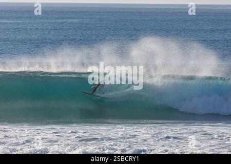 Surfer in black wetsuit braves the winter surf and rides a wave in clear blue green waters Stock Photo