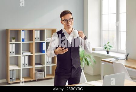 Happy young businessman standing in office, drinking coffee and using mobile phone Stock Photo