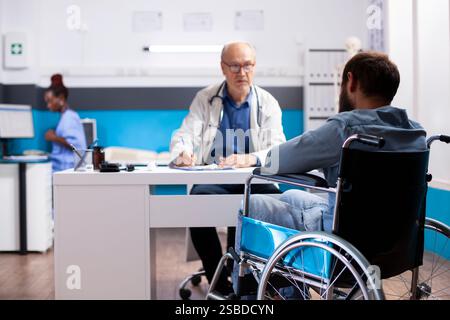 Older doctor in lab coat writing on clipboard as sick male patient mentions his symptoms in clinic. Bearded man in wheelchair speaking to senior physician about recovery progress at medical checkup. Stock Photo