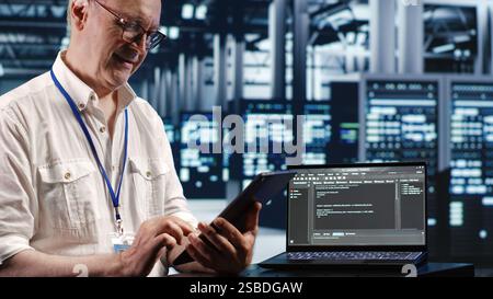 Elderly worker in data center, solving tasks on tablet while running scripts on laptop terminal. Certified engineer amidst servers, accessing databases and manipulating lines of code Stock Photo