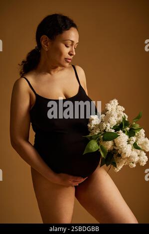 A serene pregnant woman wearing black clothing holding a bouquet of white flowers, expressing maternity beauty and motherhood. Photographed on a neutr Stock Photo