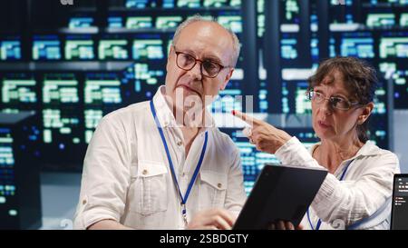 Software developers in high tech establishment, completing tasks on tablet while running scripts on laptop terminal. Proactive programmers amidst server rows, accessing databases and manipulating code Stock Photo
