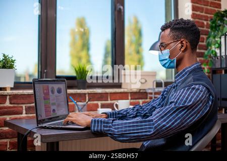 Focused manager works in startup office typing on his laptop and reviewing online documents, to improve company growth. Male project analyst with face mask and wireless earbuds, working at his desk. Stock Photo
