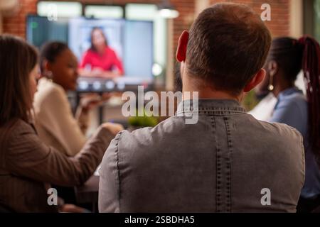 Rearview closeup of man sitting with coworkers and having online discussion with female employer on digital monitor. Male employee with beard looking at screen displaying video call of businesswoman. Stock Photo