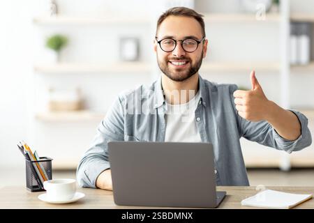 Happy young guy sitting at table with flag of Canada, using laptop computer, showing thumb up gesture at home Stock Photo