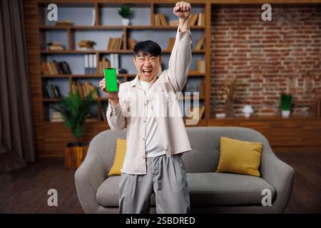 Excited surprised man, winning lottery, phone screen, surprised, asian, young man celebrating a victory while holding a smartphone in a cozy living Stock Photo