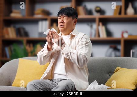 ill sick man, feeling unwell, having headache, asian man sitting on a sofa with tissues in hand, capturing a moment of sadness and reflection at home Stock Photo