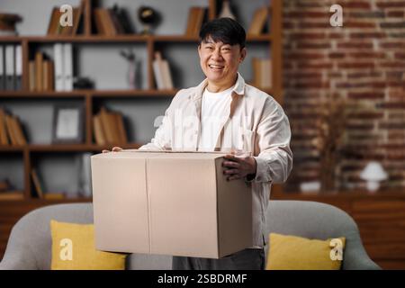 opening delivery box, excited man, Online shopping concept, Smiling asian man holds large cardboard box in cozy living room filled with books and Stock Photo