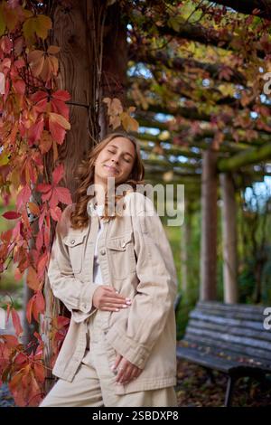 Autumn Portrait: Smiling Young Woman in Beige Outfit Leaning on Wooden Pergola with Red Leaves. Cozy Fall Scene Showcasing Natural Garden Background Stock Photo