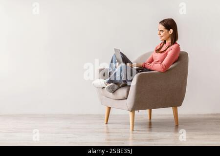 Positive young lady sitting in armchair with laptop pc, studying remotely or video conferencing against white wall Stock Photo