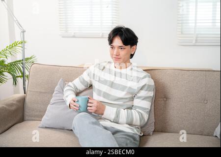 A handsome, happy young Asian man enjoys his morning coffee on the sofa in the living room while watching the news on TV. domestic life, lifestyle Stock Photo