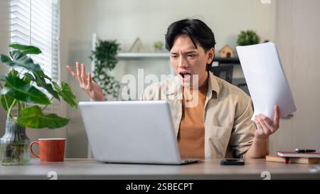 A displeased, unhappy Asian businessman sits at his desk in the office, holding reports and looking at his laptop screen with a disappointed expressio Stock Photo