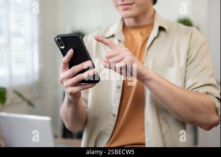 A close-up of a positive Asian man in casual casual, standing in his office, using his smartphone to text someone or read a message. people and wirele Stock Photo