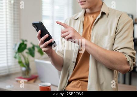 A close-up of a positive Asian man in casual casual, standing in his office, using his smartphone to text someone or read a message. people and wirele Stock Photo