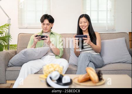 A happy young Asian couple enjoys playing a mobile game on their smartphones, spending time together on the weekend at home. domestic life, lifestyles Stock Photo