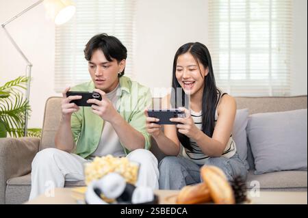 A happy young Asian couple enjoys playing a mobile game on their smartphones, spending time together on the weekend at home. domestic life, lifestyles Stock Photo