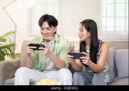 A happy young Asian couple enjoys playing a mobile game on their smartphones, spending time together on the weekend at home. domestic life, lifestyles Stock Photo