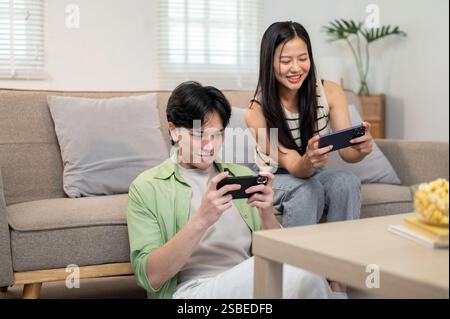 An overjoyed, happy Asian couple enjoys playing a racing game on their smartphones together, having a fun time at home. enjoyment, competition, entert Stock Photo