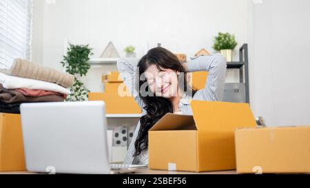 A carefree, happy Asian female online shop owner is stretching her arm to relax and relieve muscle soreness while sitting at her desk, working in her Stock Photo