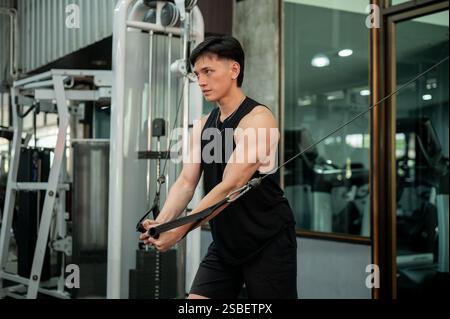 A determined and strong Asian man in sportswear is working out on a cable machine in the gym, pulling the cables to train his biceps. weight training, Stock Photo