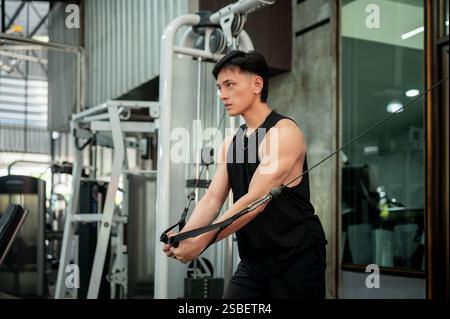 A determined and strong Asian man in sportswear is working out on a cable machine in the gym, pulling the cables to train his biceps. weight training, Stock Photo