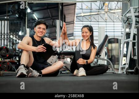 A cheerful Asian male fitness trainer and his female client are sitting on the floor in the gym, giving each other a high five after finishing their w Stock Photo
