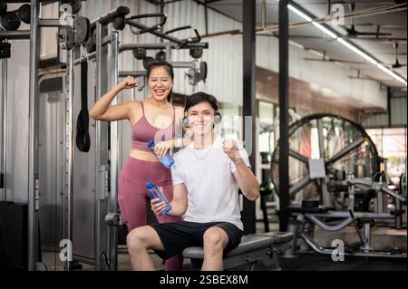 A cheerful and fit Asian male and female, both in sportswear, are smiling and showing a fist to the camera while standing in a professional gym. gym l Stock Photo