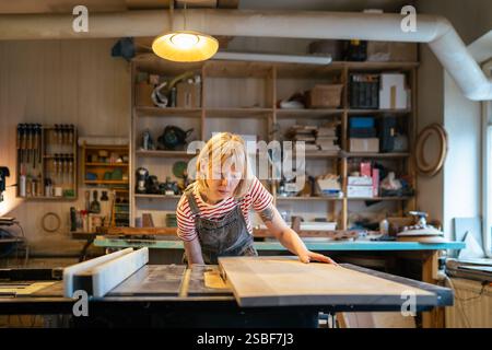Professional female joiner sets cutting depth for wood board on woodworking machine in workshop Stock Photo
