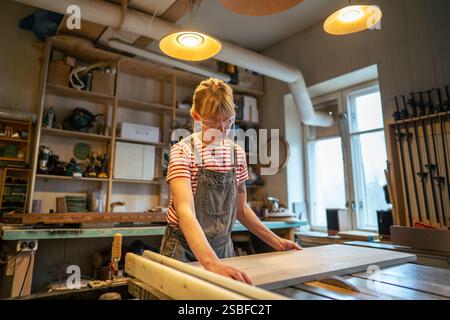 Woman carpenter in protective goggles cuts wooden piece on woodworking machine in craft workshop Stock Photo