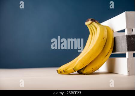 A bunch of ripe yellow bananas leans against a white wooden crate, set against a deep blue background for a clean and modern composition. Stock Photo