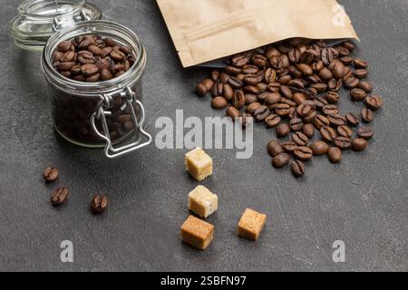 Roasted coffee beans in glass jar and in paper bag. Pieces of brown sugar on table. Black background. Top view. Stock Photo