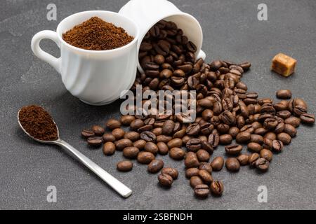 Ground coffee in cup and spoon. Roasted coffee beans are scattered from cup onto table. Black background. Close up. Stock Photo