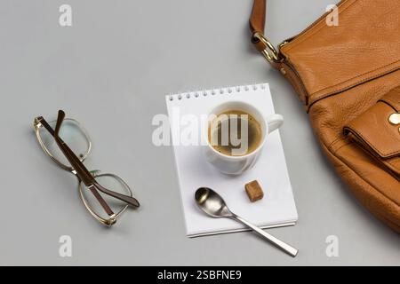 Cup of coffee, spoon and sugar cubes on notebook. Glasses, postcard and brown handbag on table. Gray background. Top view. Stock Photo