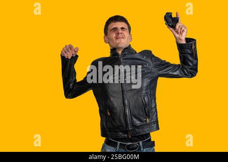 Young man wearing black leather jacket holding broken controller celebrating victory on yellow background Stock Photo