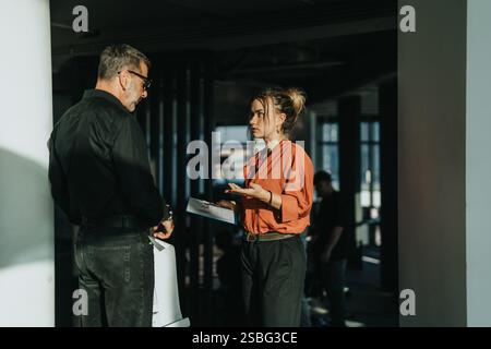 Two business people engage in a focused conversation in a contemporary office setting, exchanging ideas and holding documents. Stock Photo