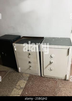 The image showcases three metallic safes, two white and one black, positioned against a plain white wall. The safes are placed on a textured stone flo Stock Photo